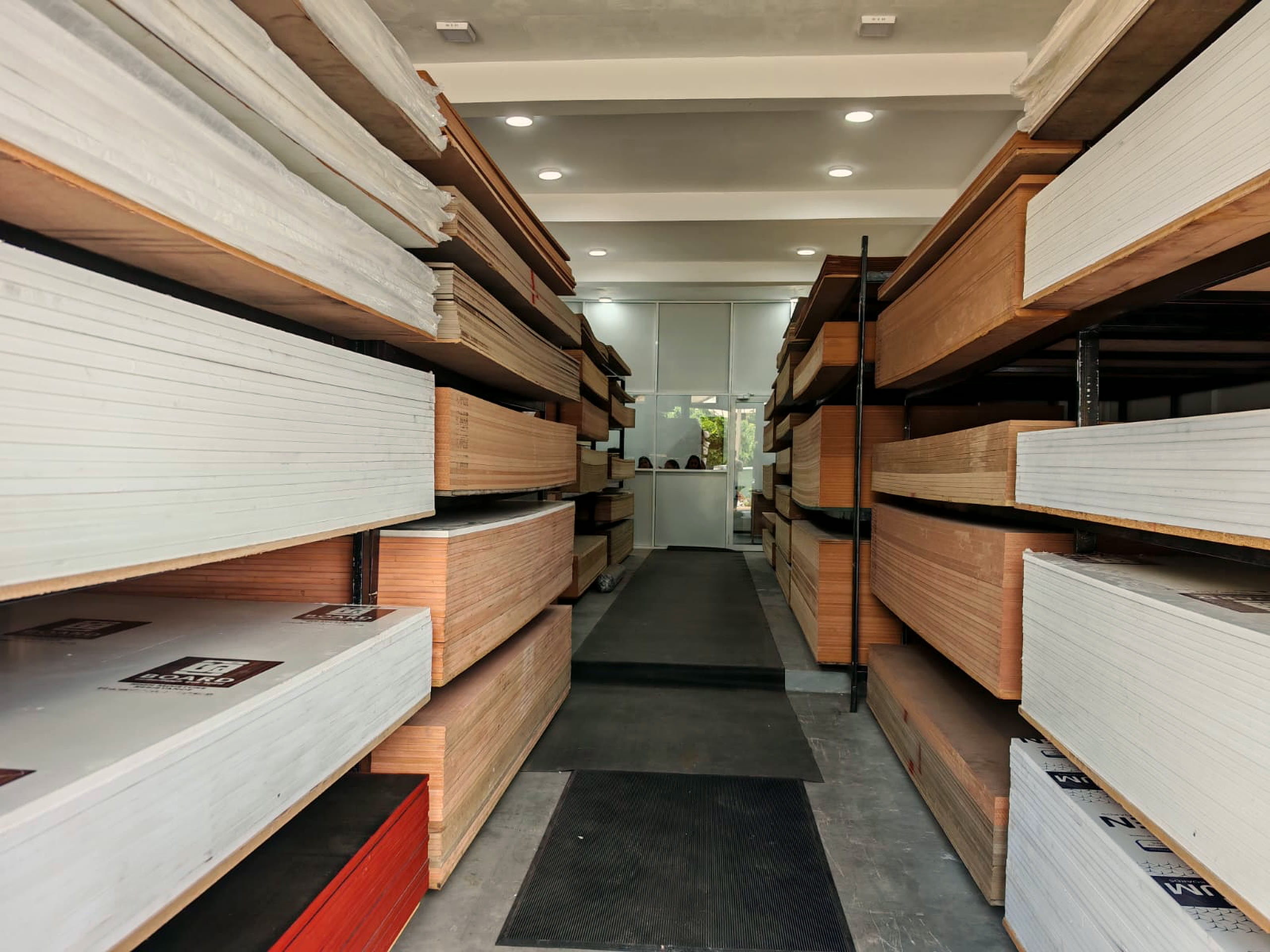 The Greenply Agencies showroom aisle in Piliyandala, with plywood, MDF and melamine boards stacked on black steel racking either side, leading to the glass fronted counter.