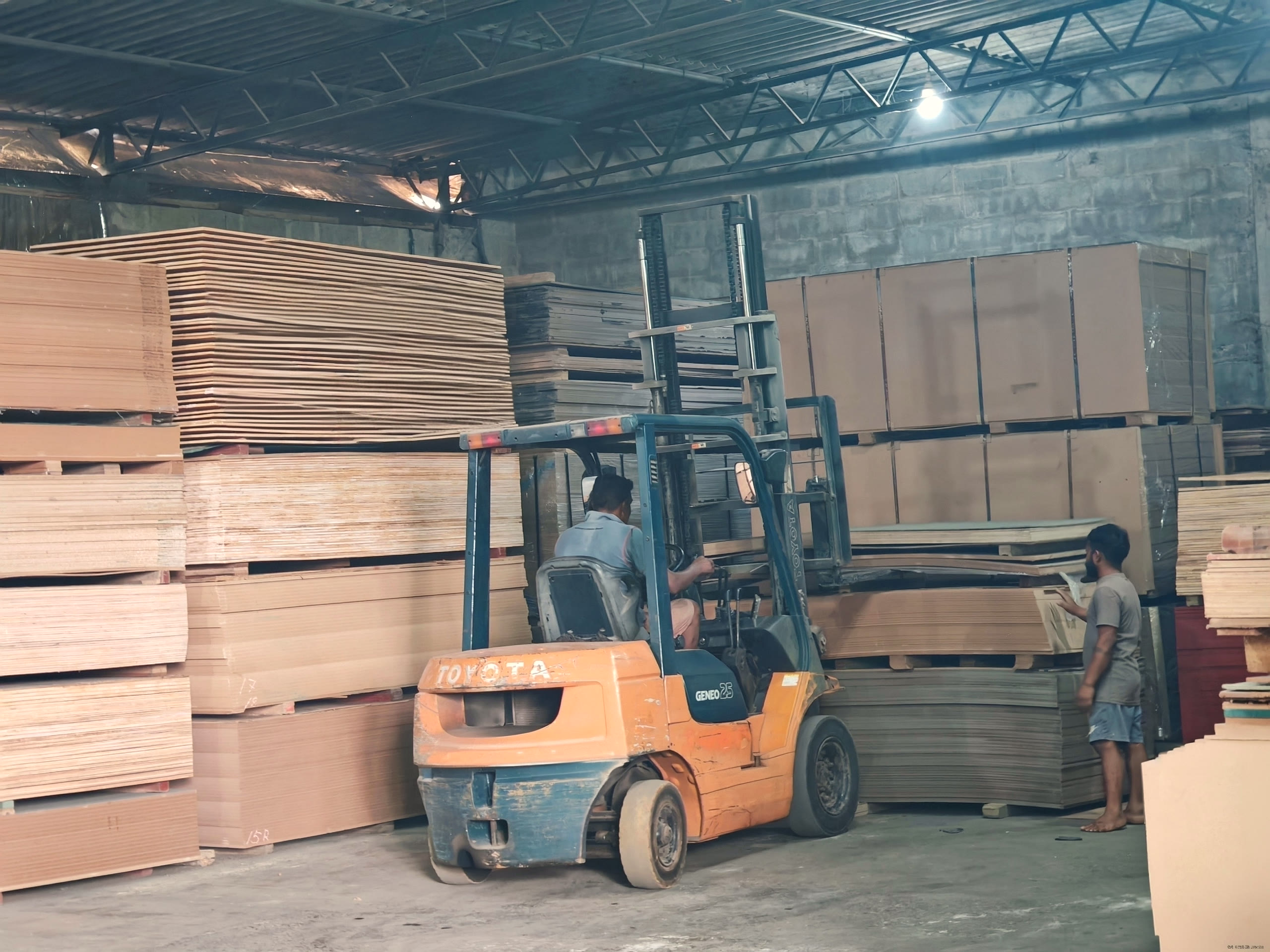 The warehouse floor with stacked pallets of boards along the walls and a forklift working on the right.